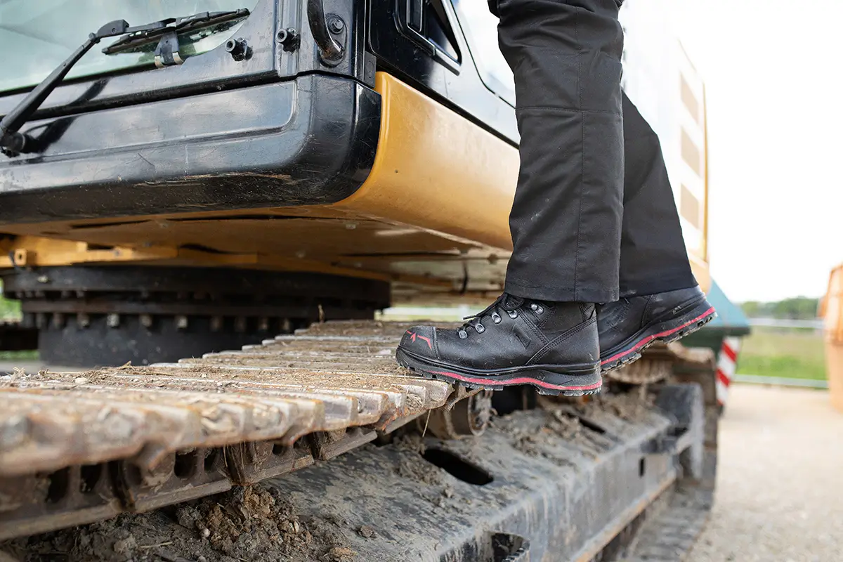 HAIX Trekker Pro 2.0 S3 Mid Sicherheitsschuh in Schwarz-Rot neben der Laufkette eines Baggers
