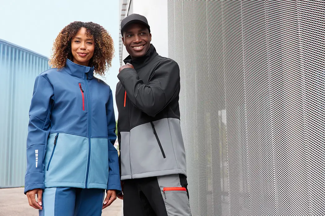 Daiber Workwear Softshelljacke JN1886 Unisex in blau/stahlblau für Damen und schwarz/grau für Herren vor einer Lagerhalle präsentiert
