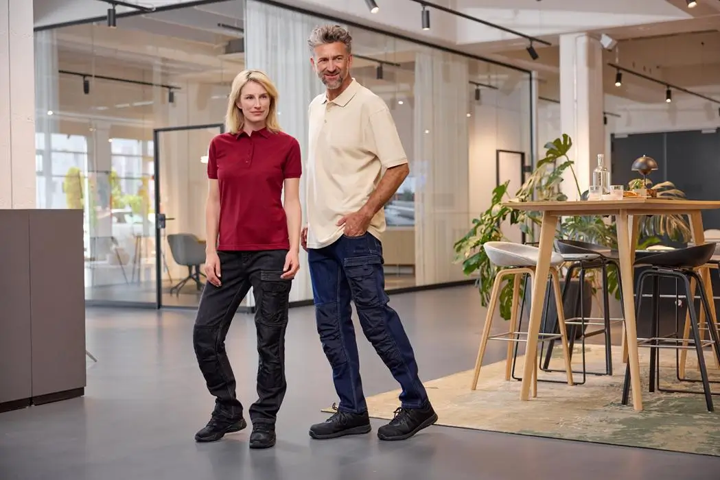 Daiber Herren Poloshirt JN874 und Daiber Poloshirt JN873 Damen Mann trägt Poloshirt JN873 in stone und Frau Mit Poloshirt JN874 in Farbe weinrot stehen in einem Raum, Lifestyleaufnahme