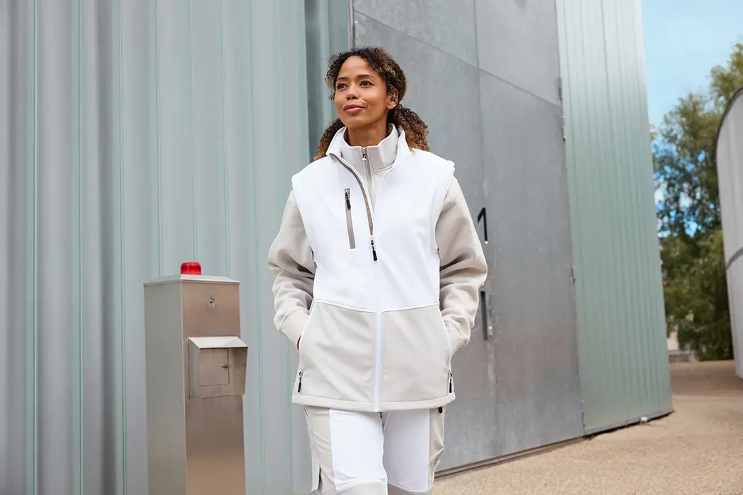 Frau trägt die Daiber Workwear Softshellweste JN1888 in white/cement vor einer Lagerhalle