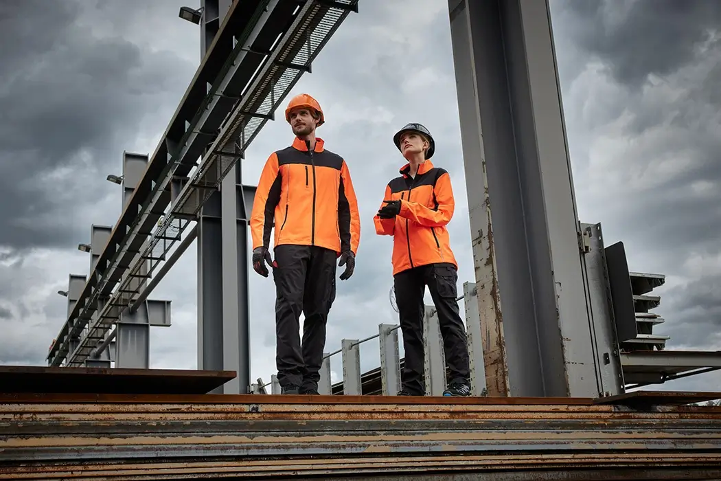 Daiber Signaljacke JN1854 neonorange/schwarz Aktionsbild von einem Mann und einer Frau auf einer Baustelle, beide tragen die neon-orange Arbeitsjacke