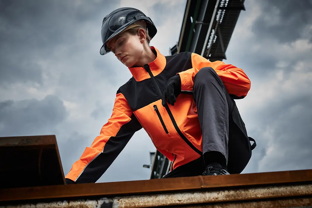 Handwerkerin auf der Baustelle, trägt eine auffälige Arbeitsjacke in neonorange-schwarz bei der Arbeit