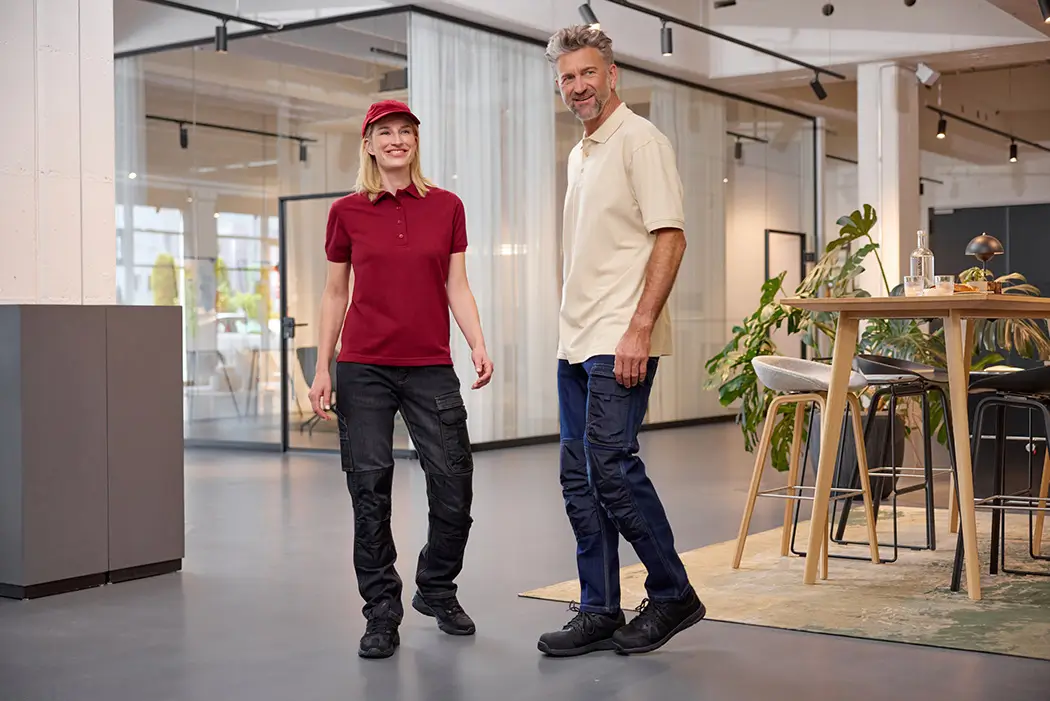 Daiber Damen Poloshirt JN873 und Daiber Herren Poloshirt JN874 Frau trägt weinrotes Poloshirt JN873 und Mann Mit Poloshirt JN874 in Farbe stone stehen in einem Raum, Lifestyleaufnahme