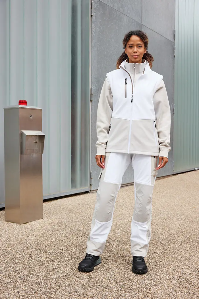 Frau trägt die Daiber Workwear Softshellweste JN1888 in white/cement vor einer Lagerhalle